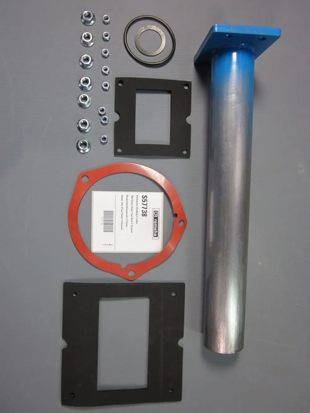 Remeha Zubehörset mit einem Metallrohr, Dichtungen, Unterlegscheiben und Schrauben, angeordnet auf grauer Unterlage.